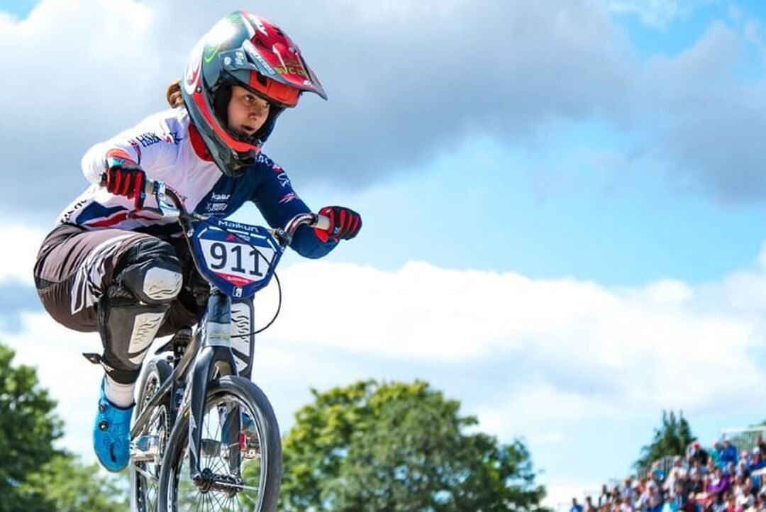 A cyclist in full gear is in the air on their BMX bike. An audience can be seen in the background.