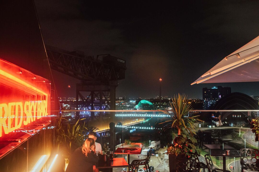 Slightly distorted view from a rooftop at night. Two people are sitting at tables and their is a neon red sign that reads 'Red Sky Bar'. In this distance you can see a river and a crane.