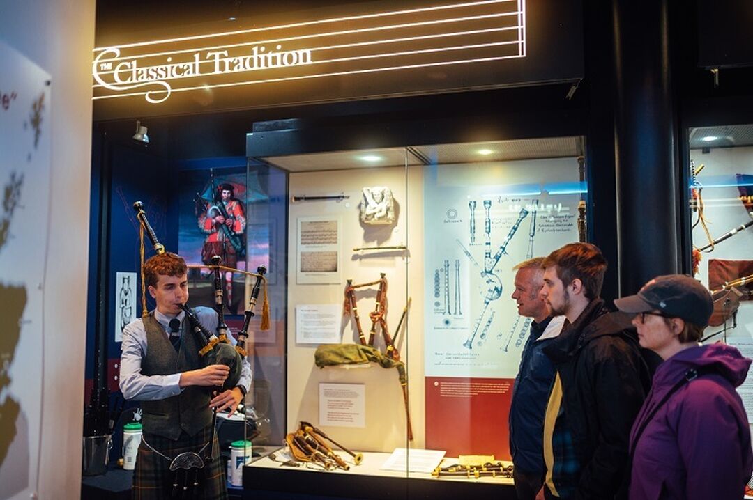 A person playing the bagpipes beside a display case full of piping paraphernalia with 3 people watching.