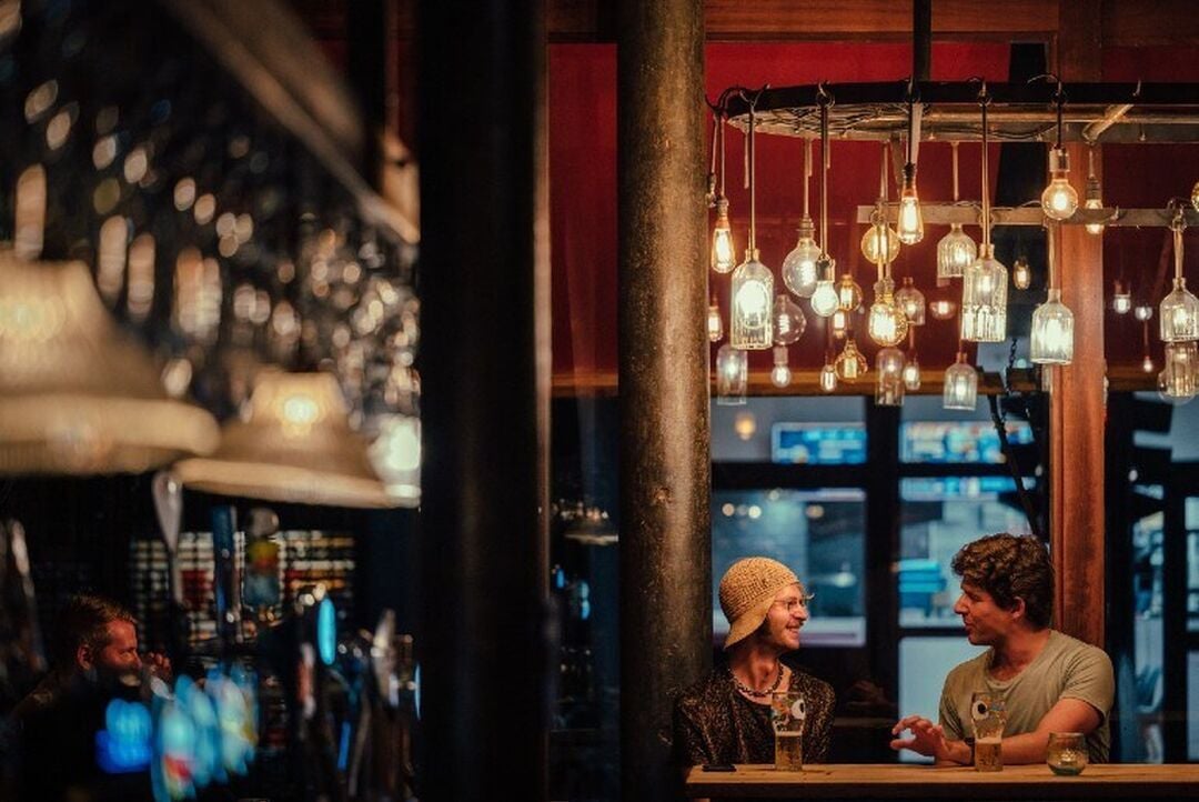 2 people sit at a bar chatting and smiling. They are under an interesting canopy of different shaped lights.