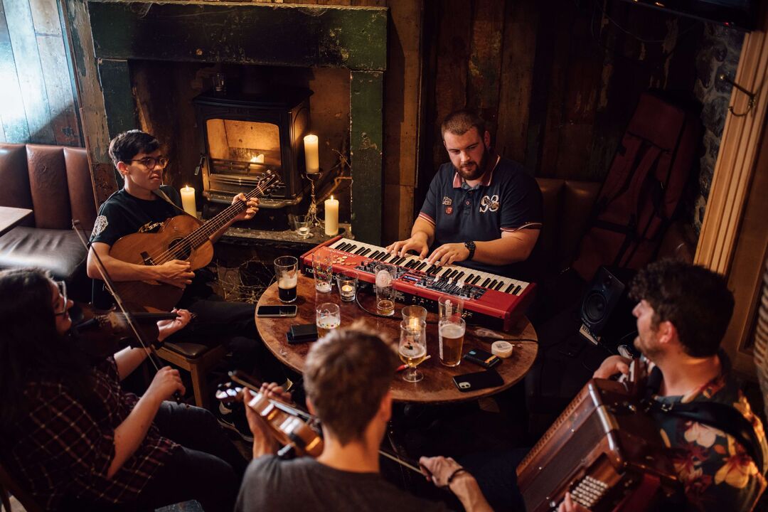 A group of musicians around a table beside a fireplace play their instruments, including a keyboard, guitar, accordion and fiddles.