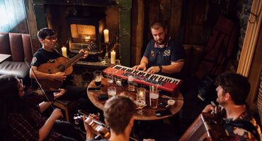 A group of musicians around a table beside a fireplace play their instruments, including a keyboard, guitar, accordion and fiddles.