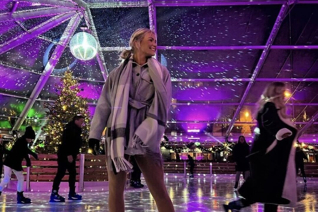 A skaker wearing a scarf smiles. A disco ball shines lights on the glass roof and ice rink.