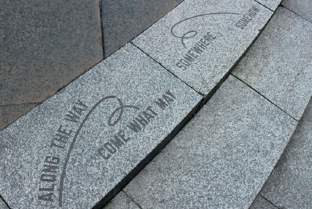 Across two paving stones are the engraved words 'Along the way, come what may, somewhere, somehow'. An engraved line swirls between the words.
