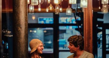 2 people chat in a pub beneath a selection of vintage style lighting.