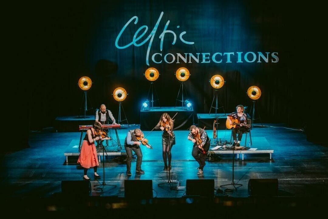 6 musicians are playing their instruments on stage against a backdrop of standing lamps and blue lighting. The curtain at the back of the stage says Celtic Connections.
