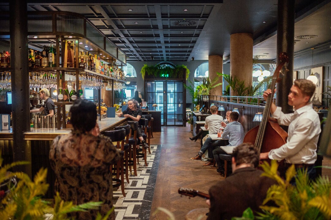A musician plays the double bass in a bar. A row of 2-seater tables beside stone pillars are on one side with a stylish bar on the other.
