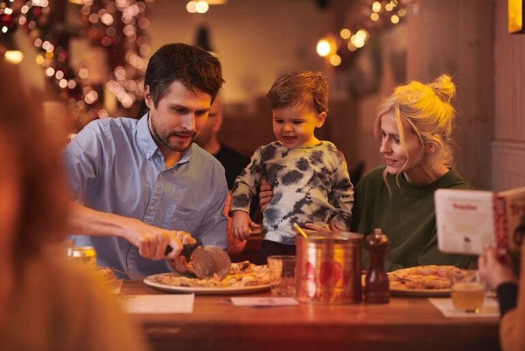 A family of 2 adults and a child sit at a table in a restaurant. One of the adults is cutting a large pizza. There are fairy lights in the background.