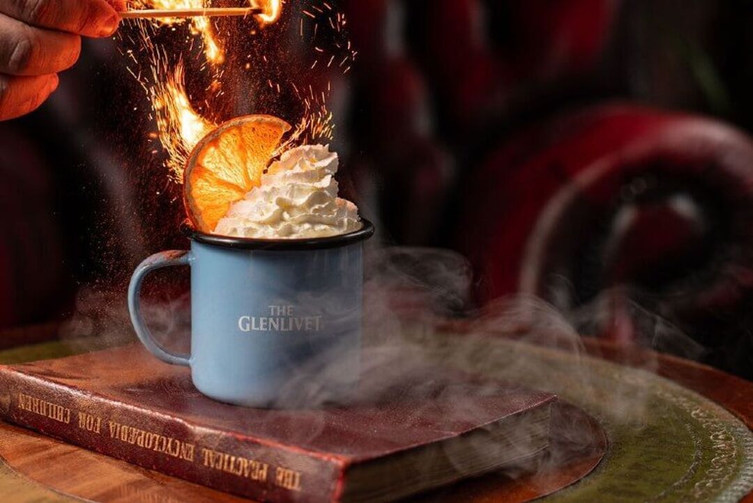 A mug topped with cream and orange peel sits on top of a book. A hand is seen with a match setting the top of the drink alight.