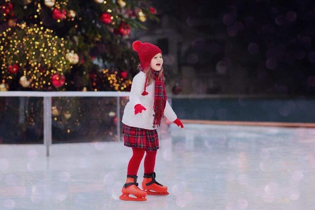 A child dressed in red and white wintery clothing skate on an ice rink in front of a large Christmas tree.