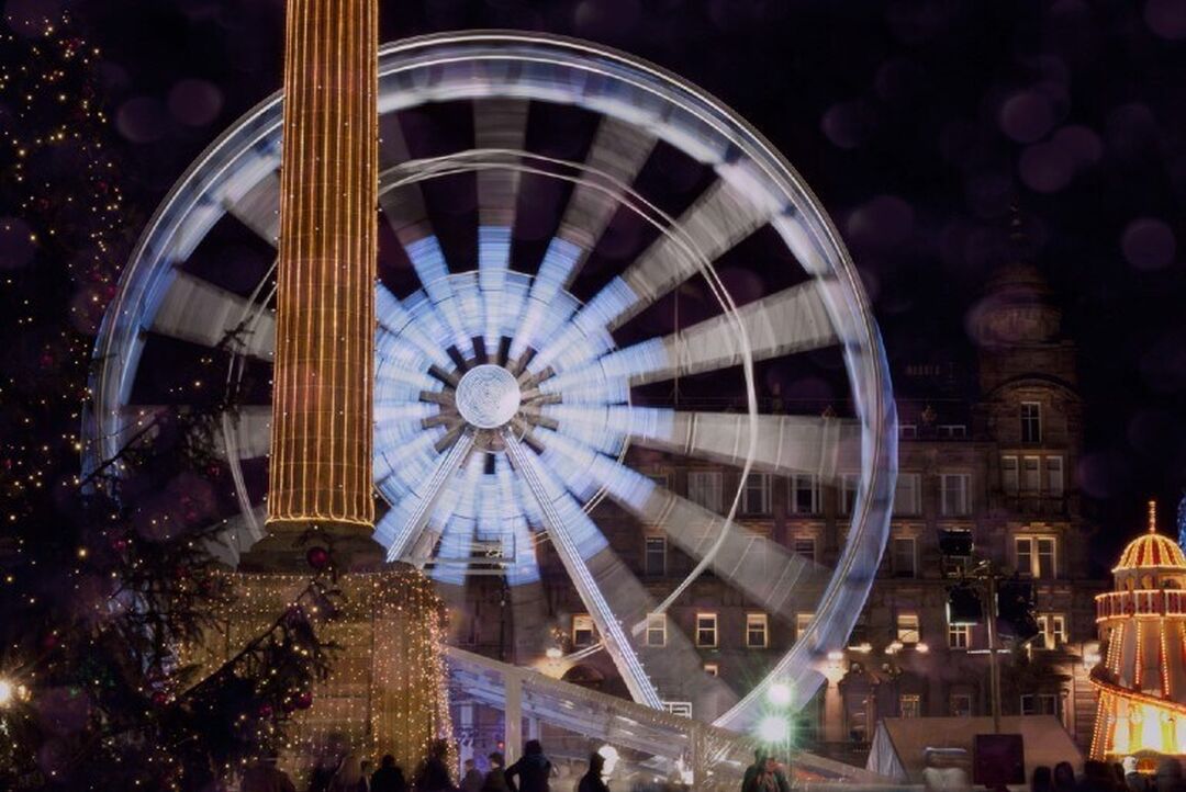 A ferris wheel lit up against the evening sky is slightly blurred. While a stone column surrounded with fairy lights in the foreground is in focus.