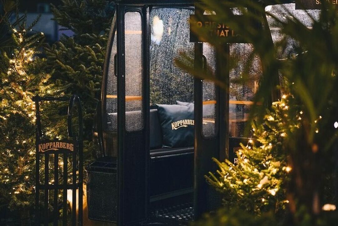 An outdoor glass booth with cushions sits surrounded by Christmas trees and fairylights.