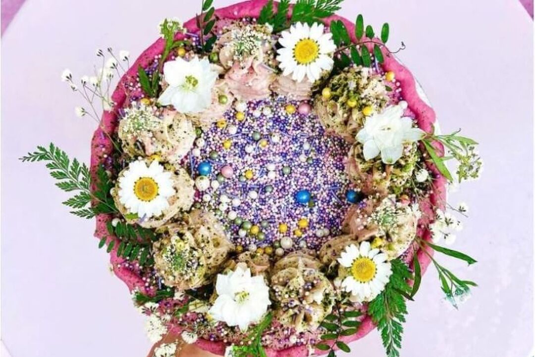 From above a colourful round raw vegan cake with pink icing, purple sprinkles and lots of real flower decorations