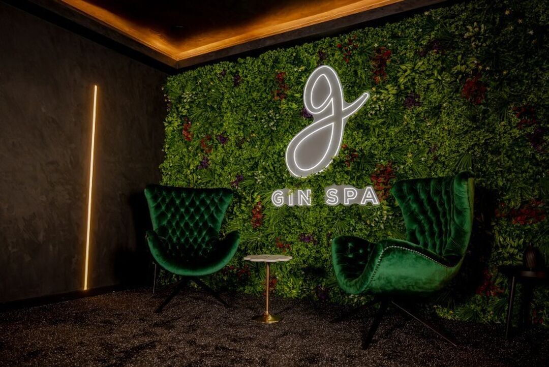 2 plush green velvet armchairs sit in front of a wall covered in greenery and with a sign saying 'gin spa'. The room is darkly lit.