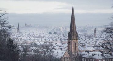 A few church spires are dotted across a city view with snowy rooftops and snowy hills in the distance.