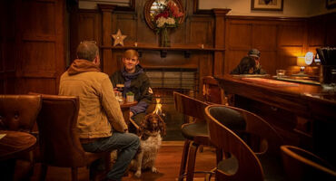 2 friends sit at a table talking with a dog by their side. A fireplace on a wooden panelled wall is behind them. Low level lighting and the fire lights the room.