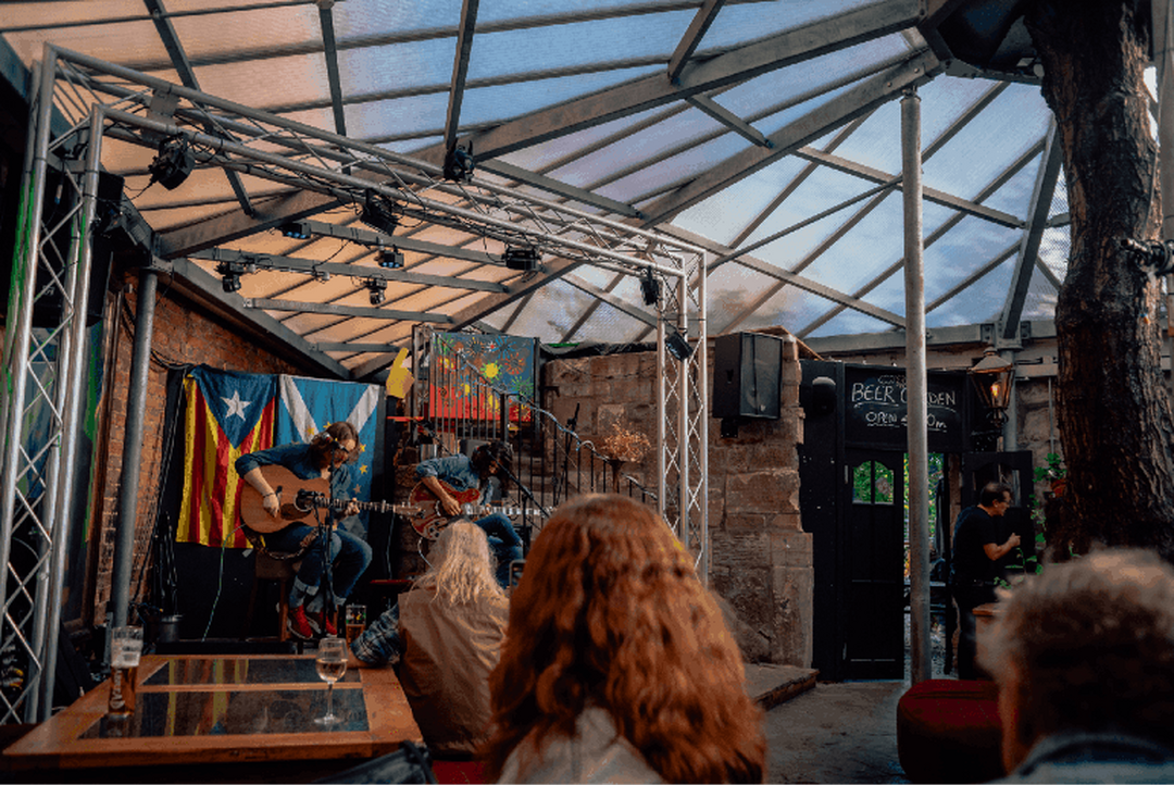 A bar with a glass panelled roof and exposed brick walls. People are sitting watching 2 musicians play guitar on a small stage.