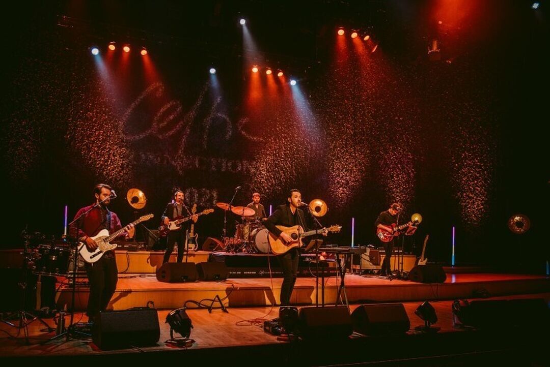 A 5-piece band perform on stage. Orange lighting lights up the stage and backdrop, which has the words Celtic Connections