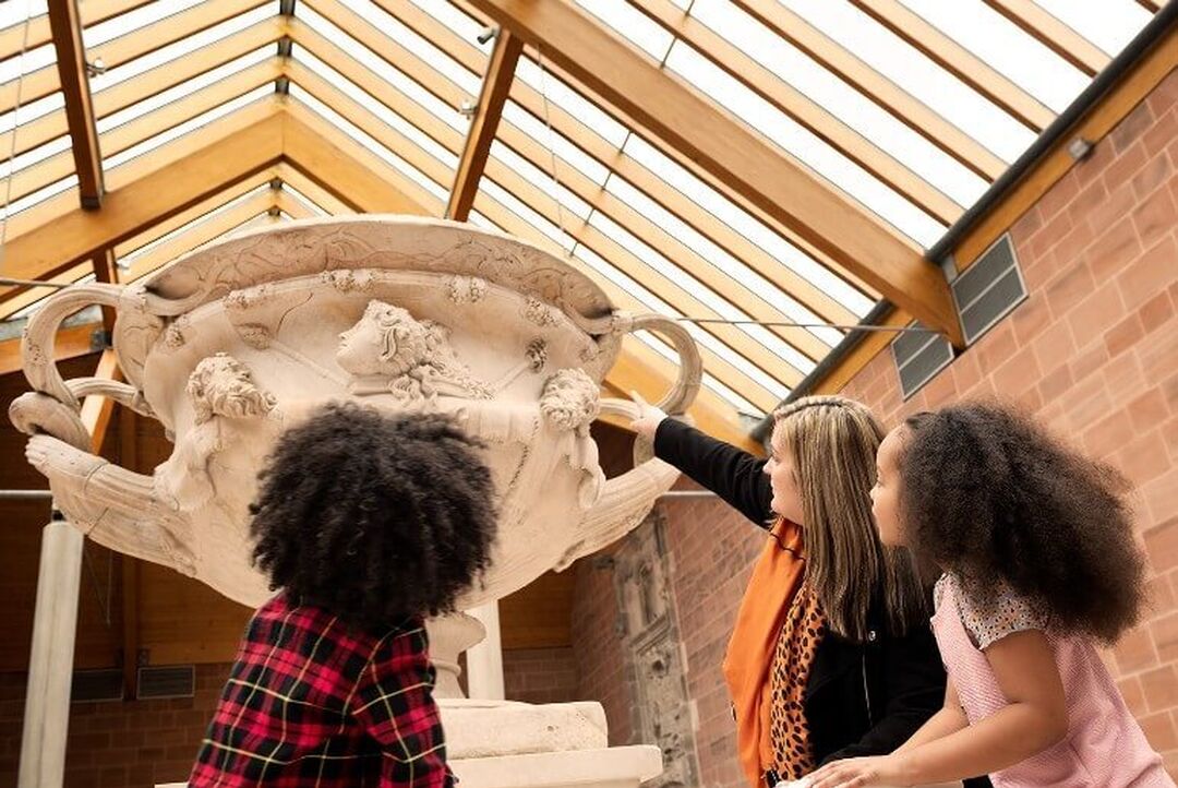 An adult points 2 children to look towards a large ceramic jug with carvings on display in a museum. The ceiling of the museum is wooden beams and windows, allowing natural light in.