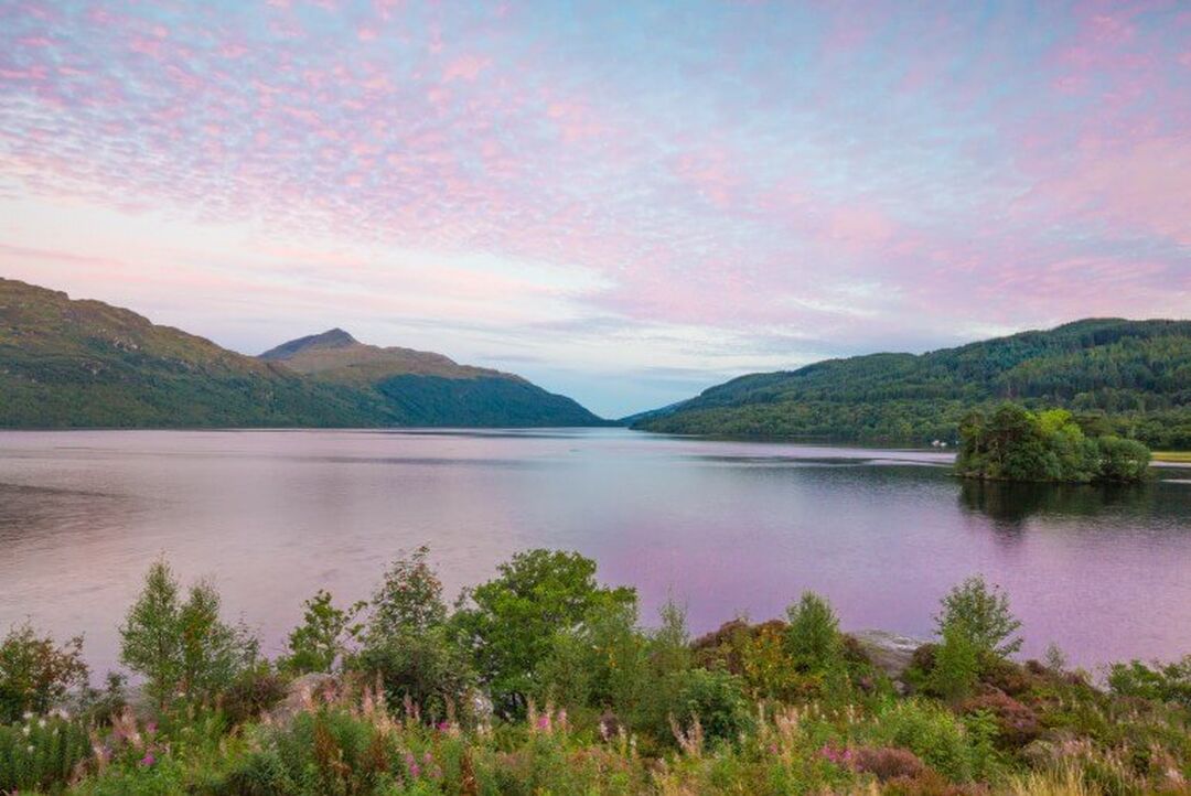 A pink sky reflects in a loch which is surrounded by undulating hills.