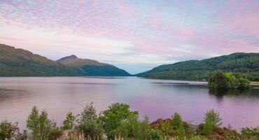 A pink sky reflects in a loch which is surrounded by undulating hills.