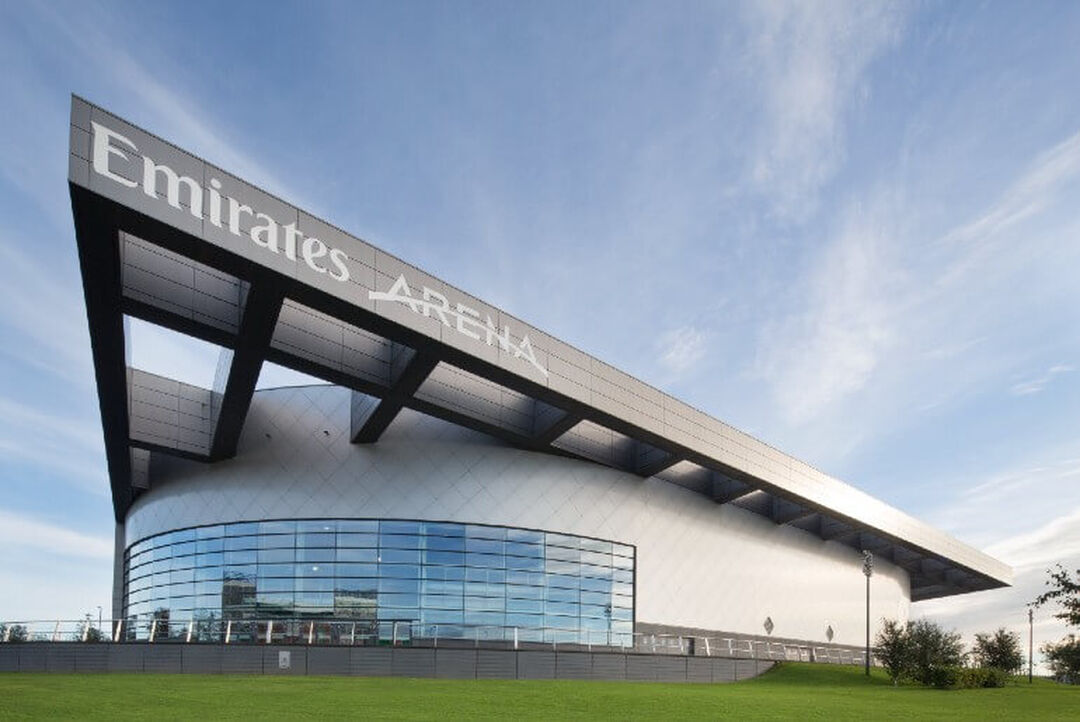 A modern silver building which has a large triangle shaped roof which says the words Emirates Arena