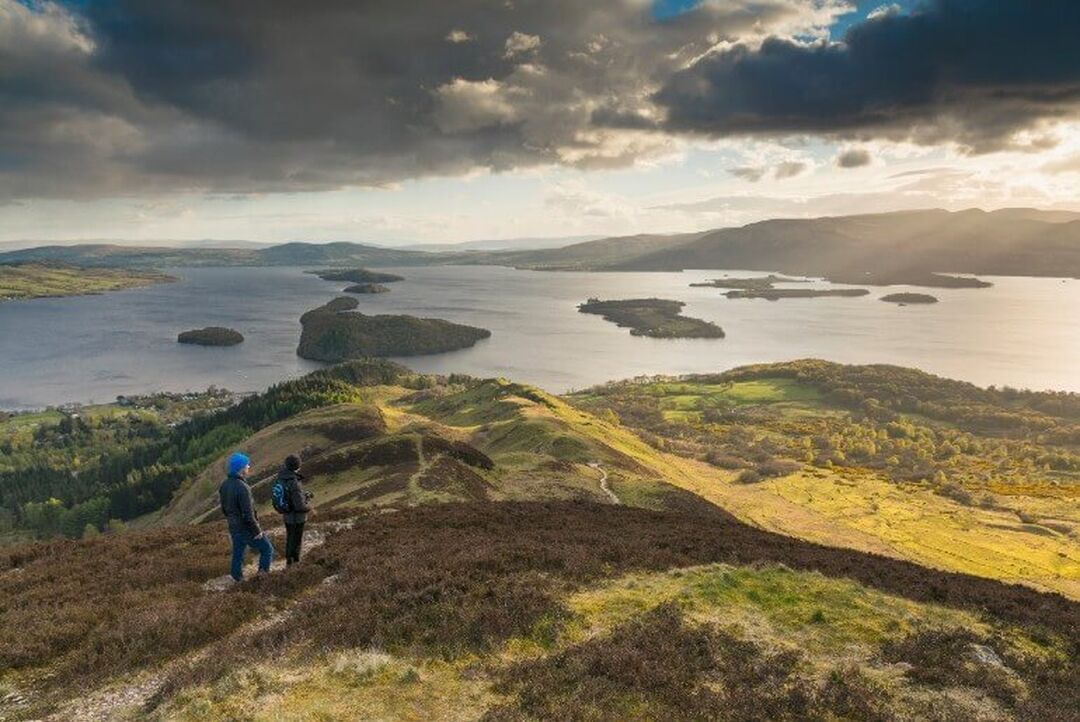2 people stand on top of a grassy hill overlooking Loch Lomond and undulating hills.