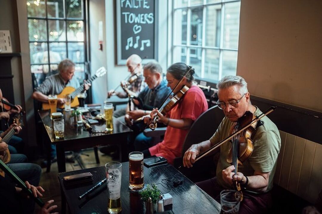 A group of musicians in a bar sit around a table with pints and play a range of instruments, including fiddles and guitars.