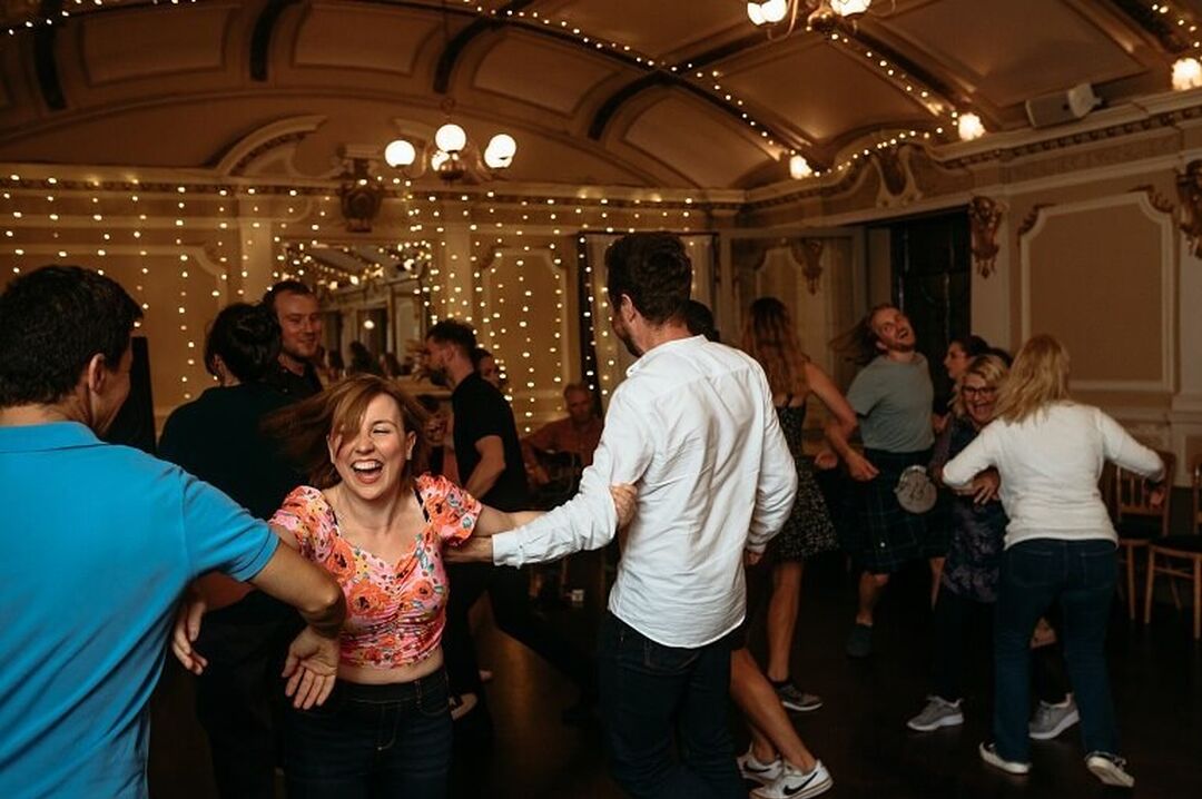 People are ceilidh dancing, arms hooked with one another. They are smiling in a ballroom that has twinkling lights.