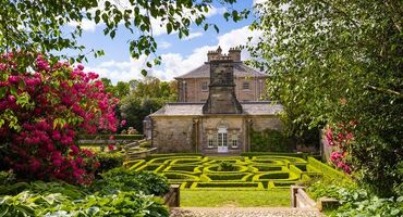 A beautiful scene of an Edwardian era mansion and its well-kept gardens including a maze and red rhododendrons in bloom.