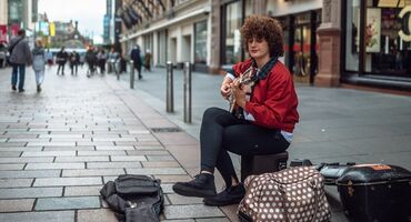 A busker sits on a cobbled pedestrianised street playing guitar with an open guitar case in front of them. Shoppers are walking on the street.