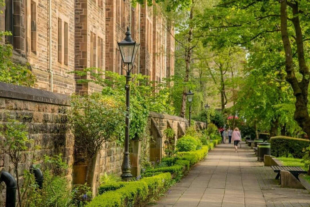 Old style lampposts, green foliage and trees line a pathway beside a historic stone building on the University of Glasgow campus.