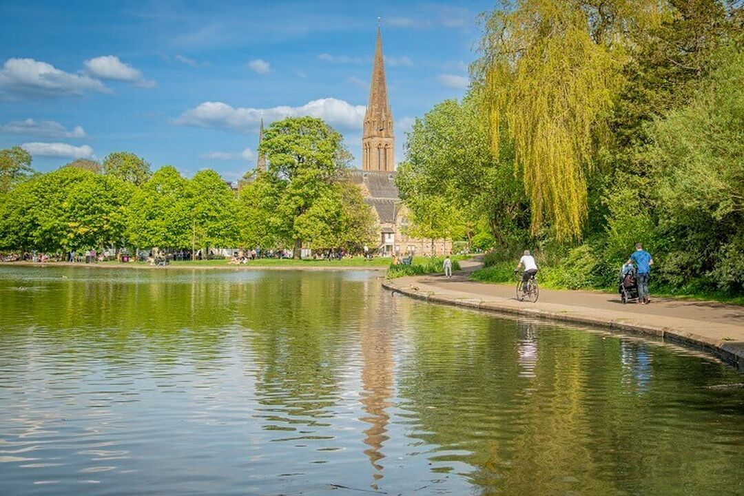 People walk and cycle around a pond in a city park on a sunny day. Trees and a church spire surround the pond.