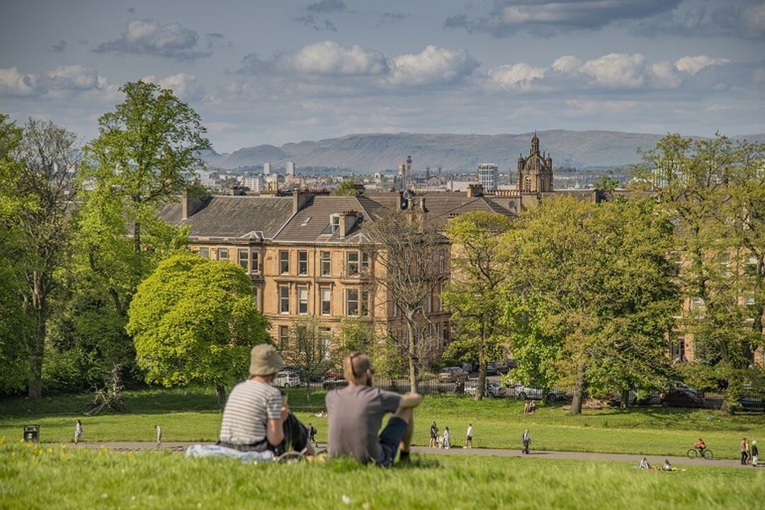 2 people sit on a grassy hill in a park overlooking a city view of houses, church spires, trees and mountains in the distance.