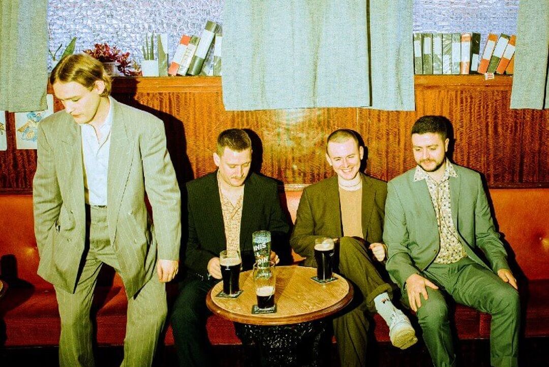 The 4 band members are laughing in a pub together with pints on the table in front of them. The style of the bar and band members is Scottish 1970s.