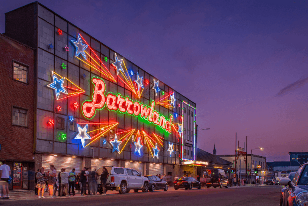 The famous Barrowland sign which says the word Barrowland surrounded by shooting stars is lit up against a dusk sky. People mingle at the front of the venue.