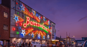 The famous Barrowland sign which says the word Barrowland surrounded by shooting stars is lit up against a dusk sky. People mingle at the front of the venue.
