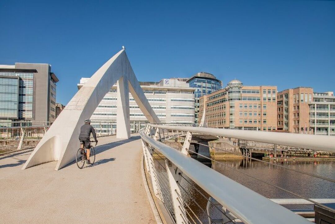 A footbridge across a river leads towards modern buildings. The bridge has a white triangular design in the centre.