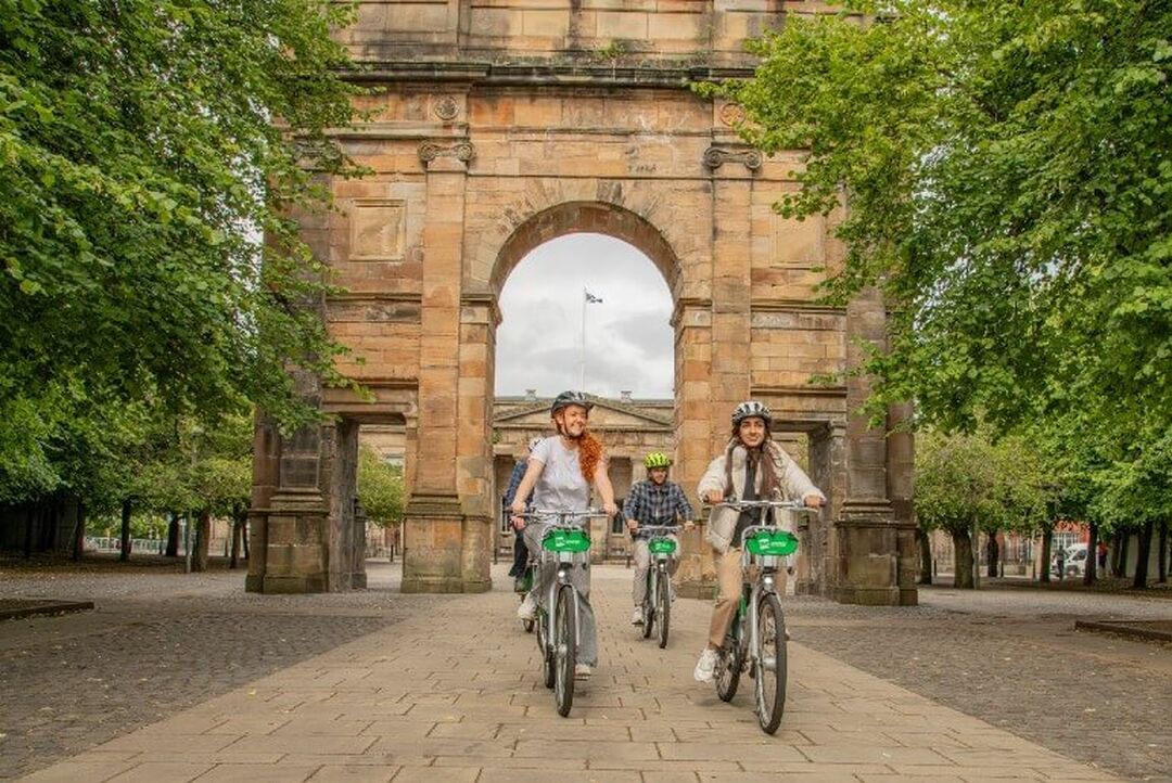 Four cyclists ride under a stone archway in a city park.