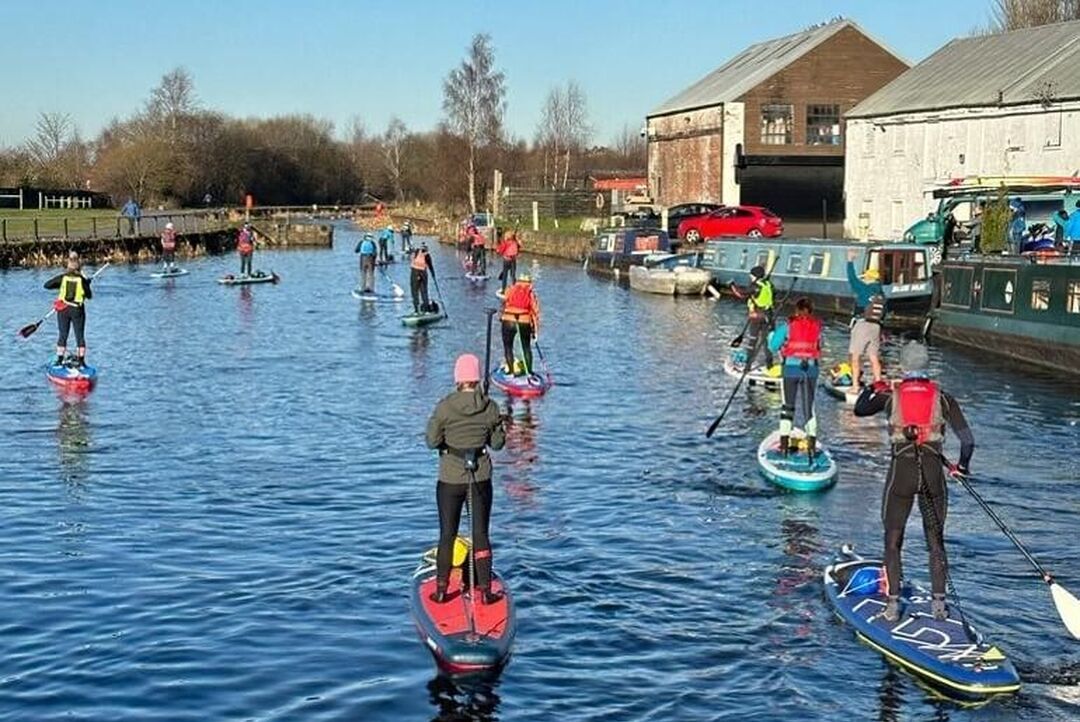 A group of paddleboarders wearing wetsuits on a canal on a sunny day.