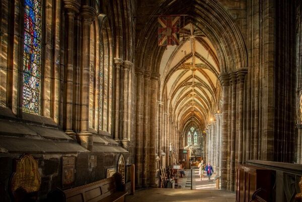 The interior of the medieval Glasgow Cathedral with stone arched walls and long thin stained glass windows. People walk beneath an archway and an old battered Union Jack flies above them.