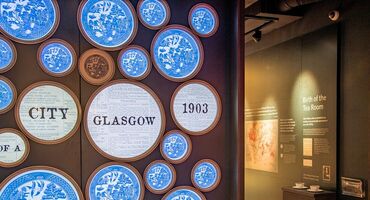 The entrance to an exhibition has a display wall of blue and white plates in a traditional Burleigh design. A few of the plates differ and read 'City of Glasgow 1903'.