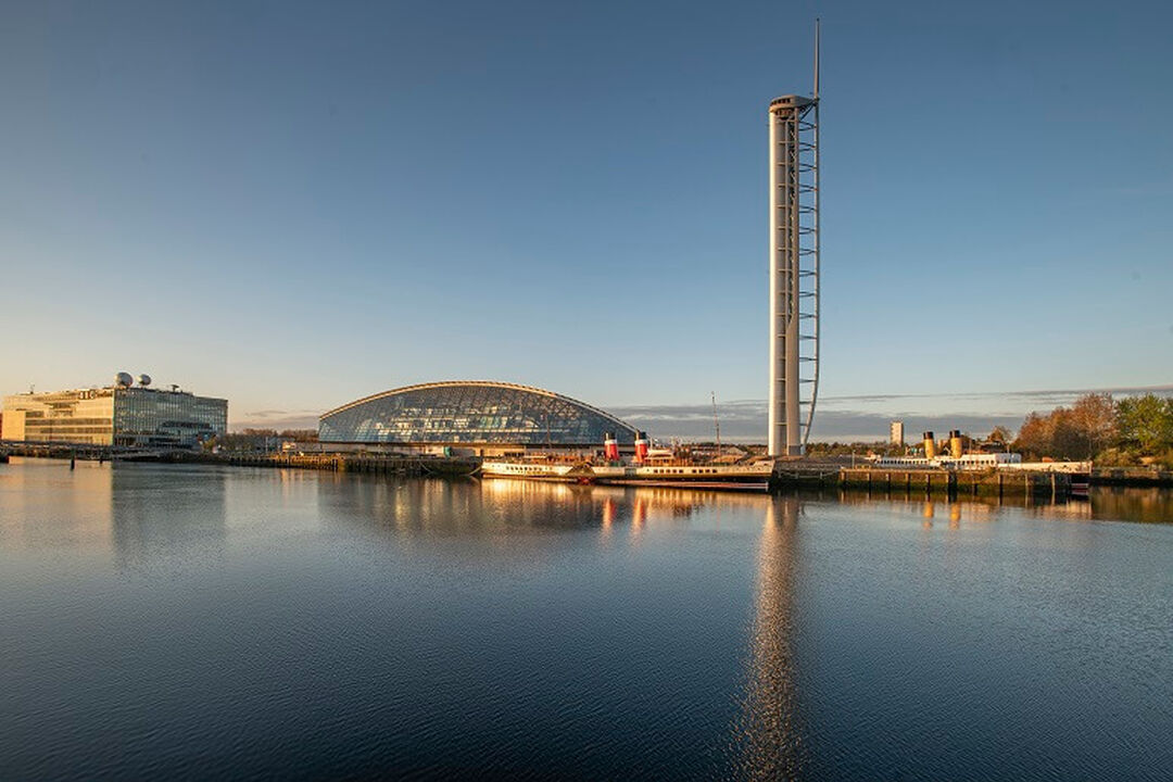A blue sky day reflects in the blue River Clyde below. The tall free standing tower and curved low building of the Science Centre is on the bank of the river.