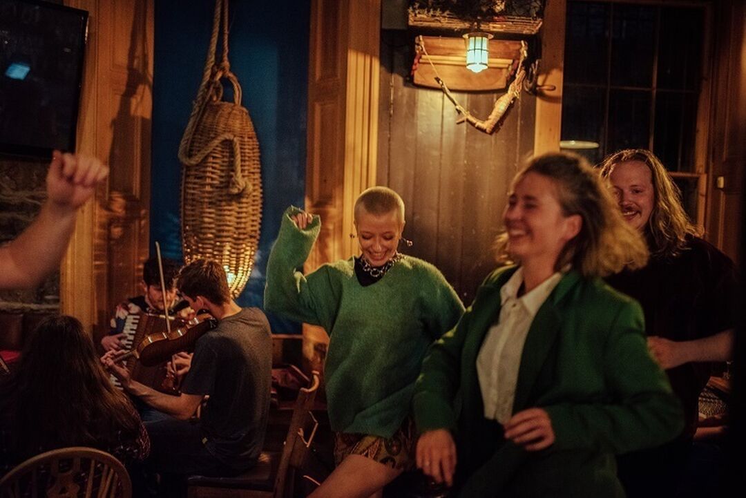 A few people smiling and dancing in a dimly lit cosy bar. Musicians can be seen jamming at a table behind them.