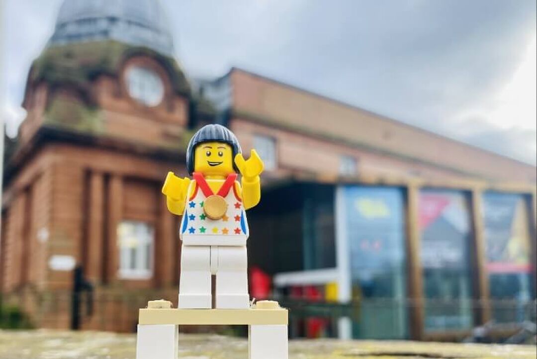A lego figure wearing a medal is in focus in front of the blurry Kelvin Hall building.