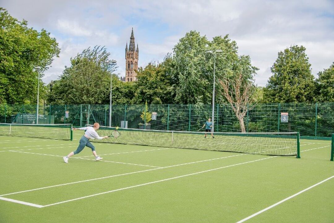 2 people play tennis on a grass outdoor court. Trees surround the court and the impressive Glasgow Uni tower can be seen between the trees.