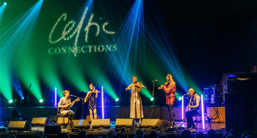 A group of musicians are on stage performing in front of a crowd. It is dark, there is green and blue strobe lighting and 'Celtic Connections' is displayed above.