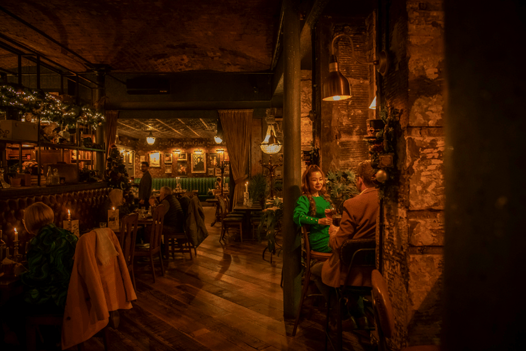 People sit at tables in a pub. Yellow lighting and candles cast a glow in the darkly lit room.
