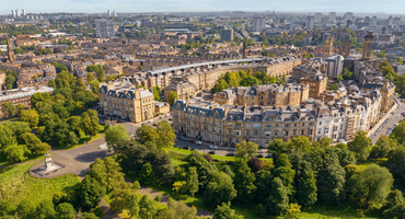 A cityscape of stone houses in a circle surrounded by green park with trees and grass, with the city in the distance.