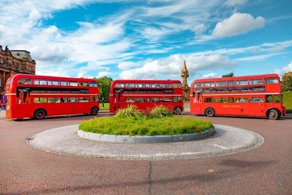 Three red old fashioned double-decker buses lined up outside two city tourist attractions, the People's Palace and Doulton Fountain.